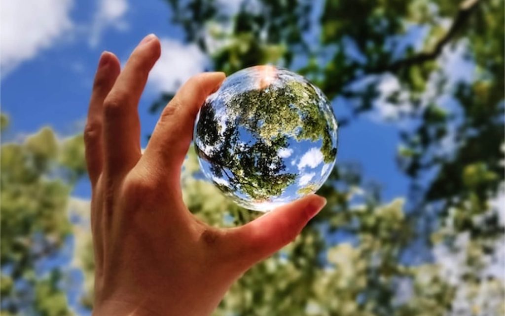 A hand holds a clear glass ball, about the size of a tennis ball, in front of a background of blue sky and trees. This makes the glass ball look like the Earth. 
