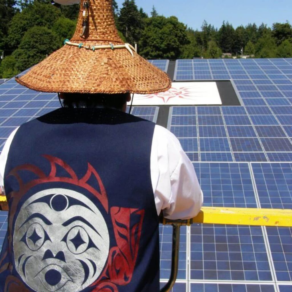 A person wearing a hat woven from wood bark and a vest with Indigenous artwork on the back looks out at a field of solar panels. 