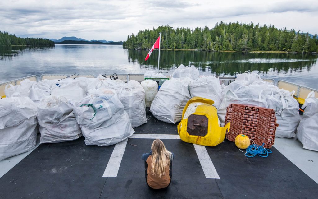 A woman with long blonde hair sits on the flat deck of a boat with a view of islands and flat water behind it. Piled in front of the woman on the deck of the boat are many white bags of waste, each of which are more than twice the woman's size. 