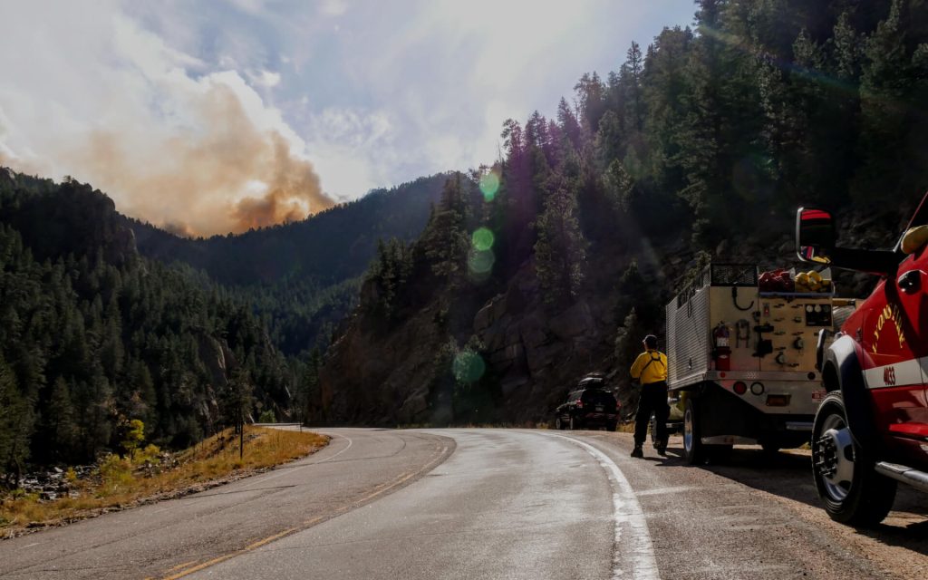 Wildfire fighter and truck on the side of the highway with a wildfire burning in the background.