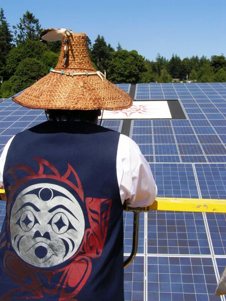 A person wearing a hat woven from wood bark and a vest with Indigenous artwork on the back looks out at a field of solar panels.