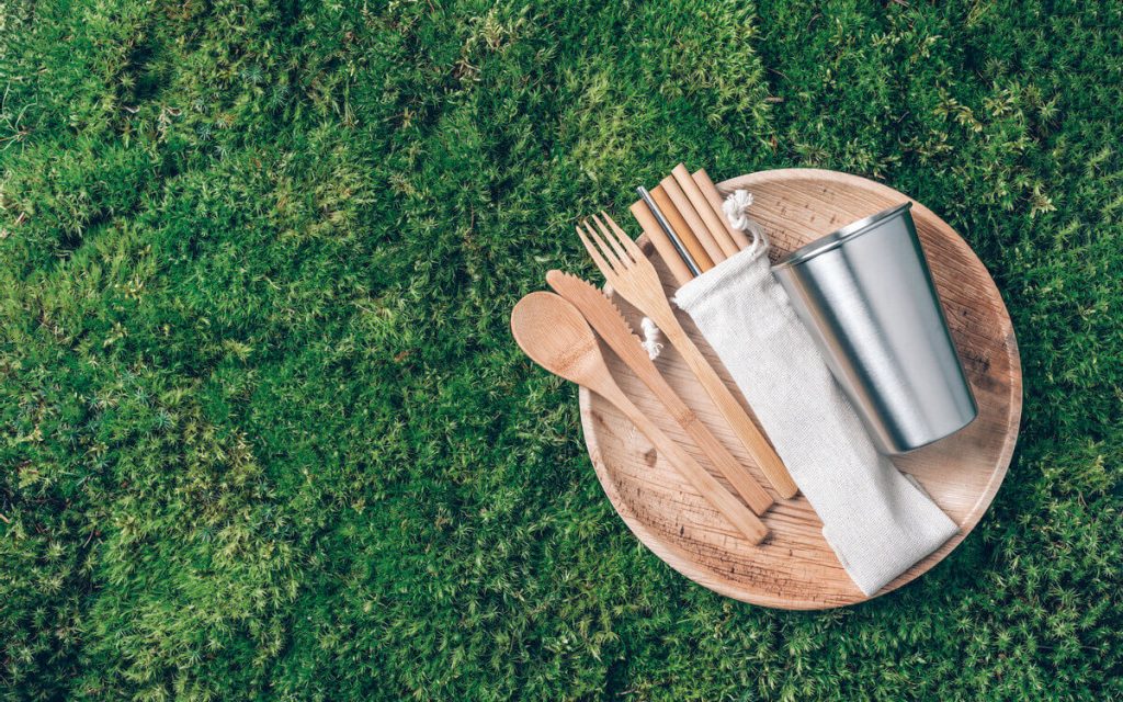 A set of wooden cutlery, paper straws and a steel tumbler on top of a wooden plate, which rests on grass.