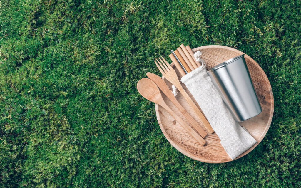 A set of wooden cutlery, paper straws and a steel tumbler on top of a wooden plate, which rests on grass.